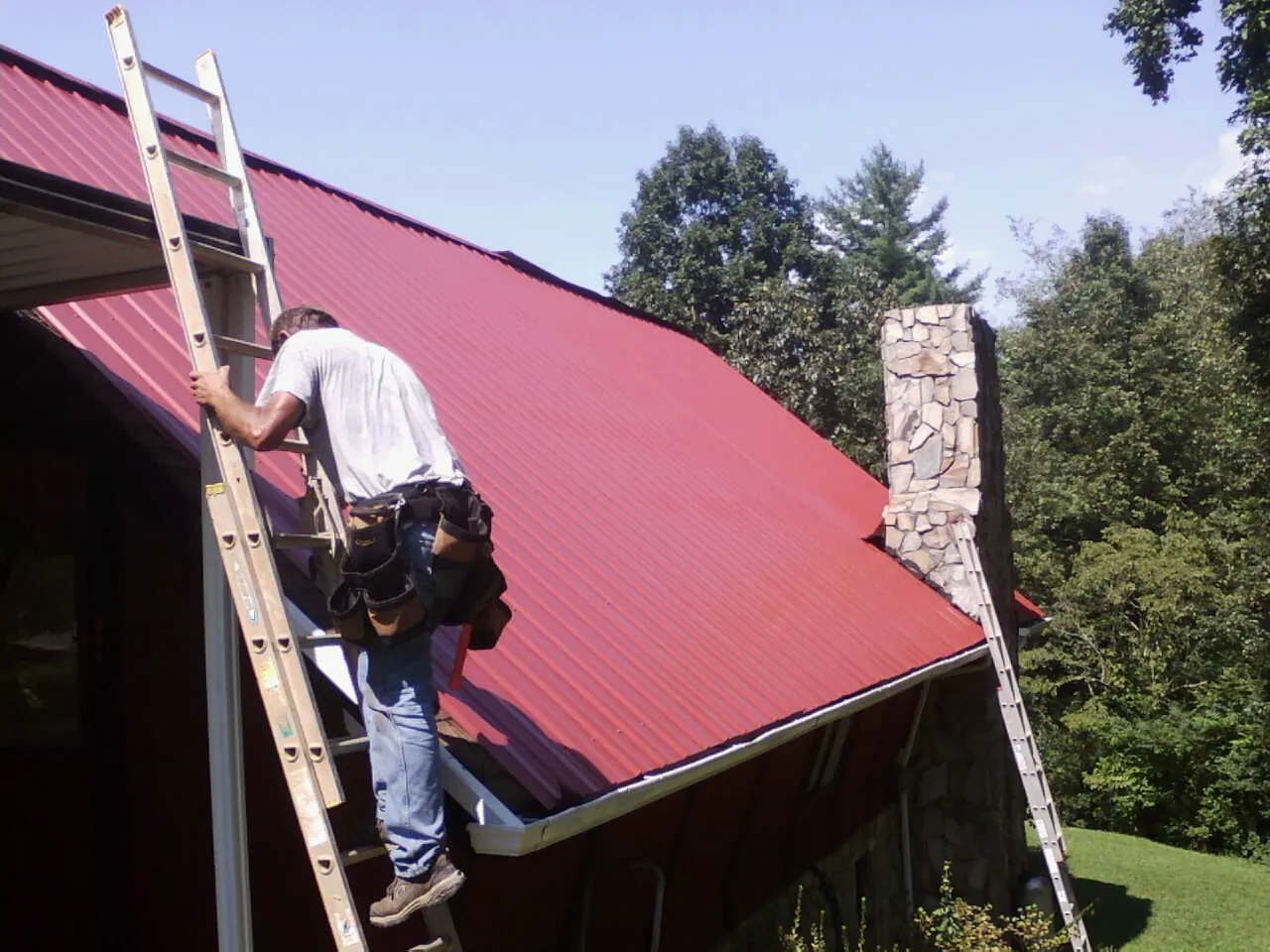 Roofer on a ladder beside a red metal roof and stone chimney — Soffit & Fascia Repair by Roofing Brothers in Valley Falls